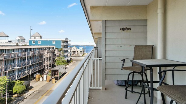 A balcony with a table and chairs, overlooking a parking area and buildings with a view of the ocean. - Salty Sands 404- 02 |Steps to Ocean & Dining (Ocean City)