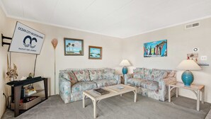 A living room with two patterned sofas, a coffee table, a side table with a lamp, and a wall-mounted sign. - Salty Sands 404- 02 |Steps to Ocean & Dining (Ocean City)