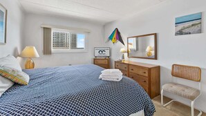 A hotel room with a bed, a dresser, a chair, a window, a mirror, and a sign. - Salty Sands 404- 02 |Steps to Ocean & Dining (Ocean City)