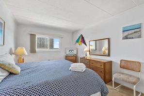 A hotel room with a bed, a dresser, a chair, a window, a mirror, and a sign. - Salty Sands 404- 02 |Steps to Ocean & Dining (Ocean City)