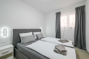 Ein Zweibettzimmer mit grauem Kopfteil, weißer Bettwäsche und grauen Vorhängen.