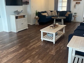Ein Wohnzimmer mit dunklem Holzboden, einem blauen Sofa, einem weißen Couchtisch und einem an der Wand befestigten Fernseher.