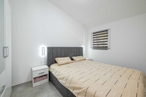 Un dormitorio con cama, mesitas de noche, ventana con persianas y una lámpara fijada a la pared.