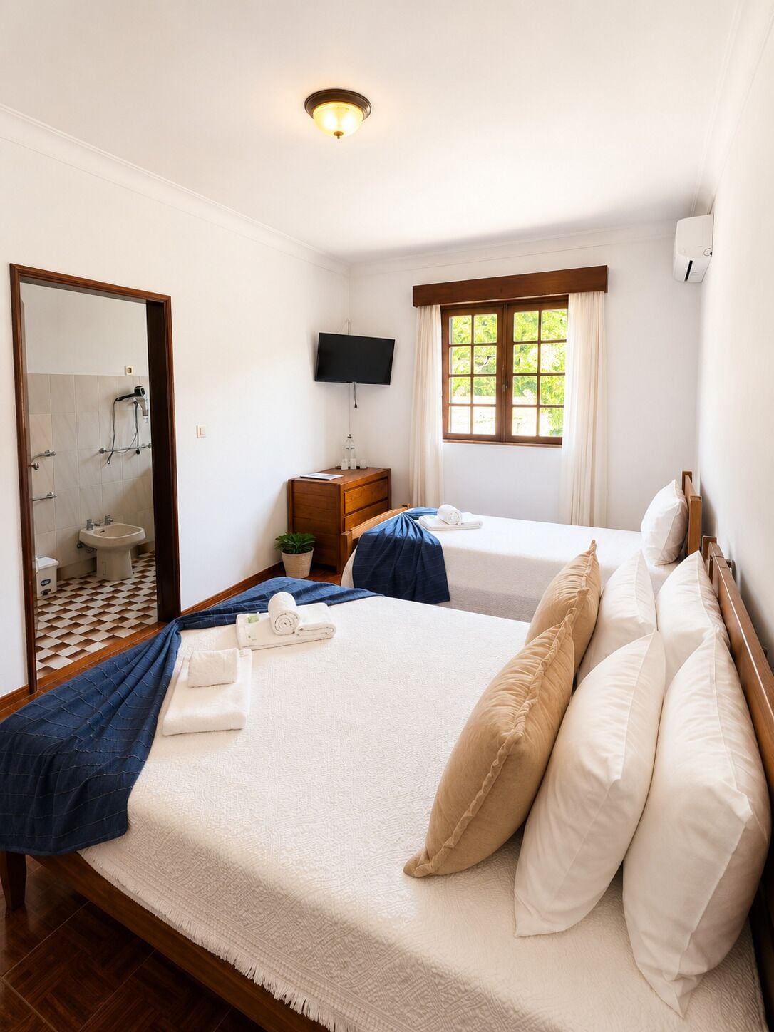 A hotel room with a bed, a TV, a window, a mirror, and a bathroom visible through the door.
