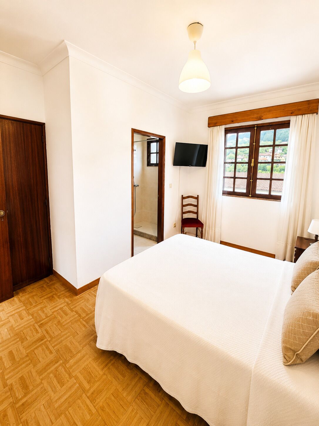 A bedroom with a bed, a TV, a window with curtains, a wooden door, and a mirror.