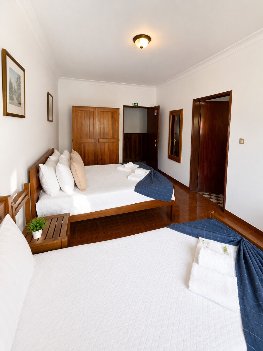 Ein Hotelzimmer mit einem großen Bett, einem Holzschrank, einem kleinen Nachttisch mit Pflanze und einer Tür zu einem weiteren Raum.