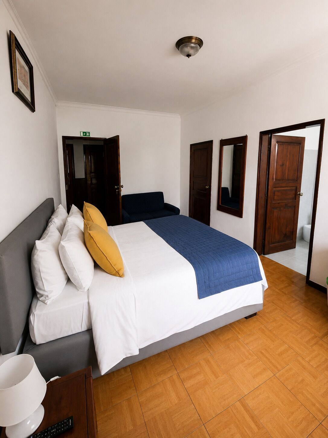 A hotel room with a bed, a bedside table, a lamp, a mirror, and a door.
