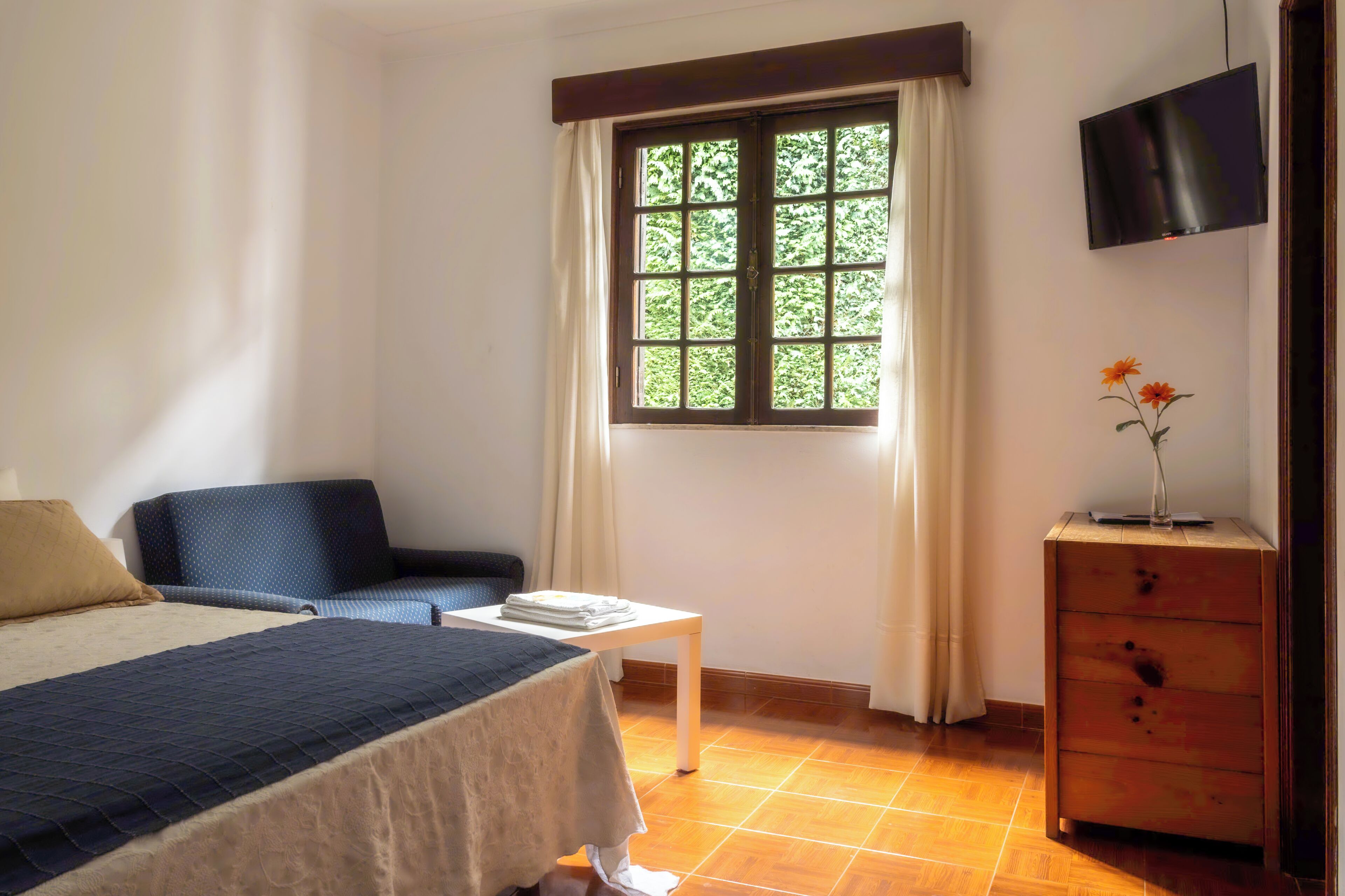 A bedroom with a bed, a chair, a small table, a TV, and a window with curtains.