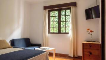 A bedroom with a bed, a chair, a small table, a TV, and a window with curtains.