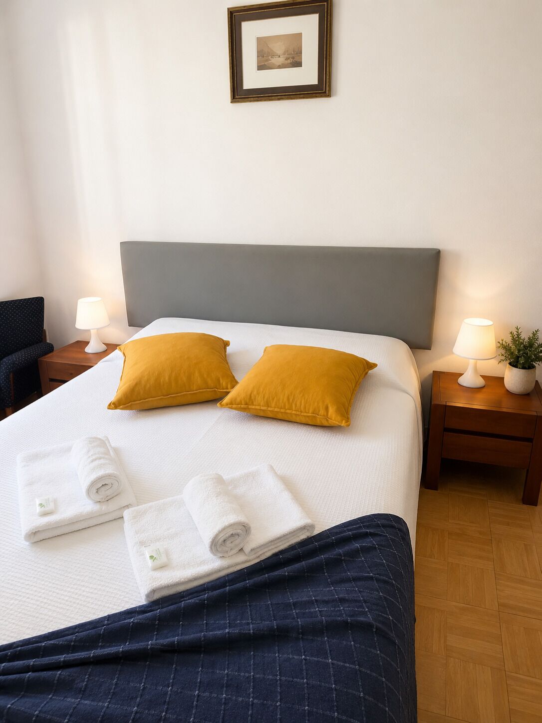 A neatly made bed with two pillows, a bedside lamp, a framed picture on the wall, and a small plant on the nightstand.