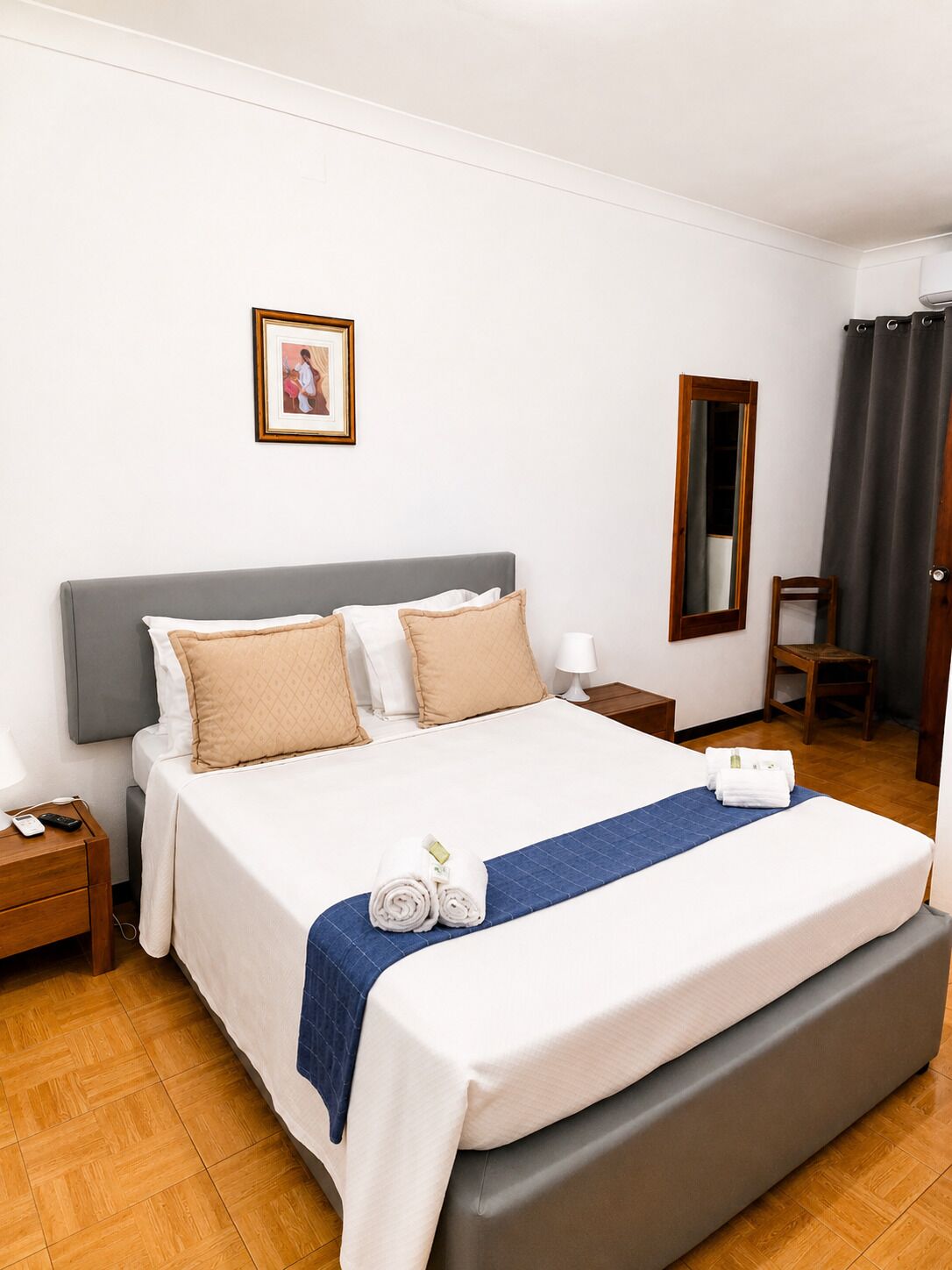 Habitación de hotel con una cama grande, una mesita de noche, un espejo, una silla y un cuadro en la pared.