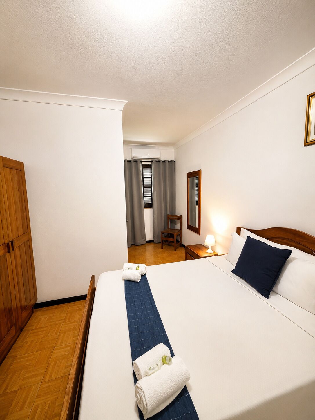 Una habitación de hotel con cama, armario de madera, silla, escritorio, espejo y ventana con cortinas.