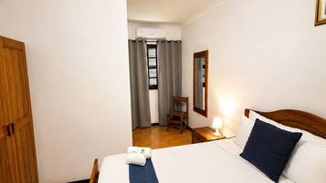 A hotel room with a bed, a wooden wardrobe, a chair, a desk, a mirror, and a window with curtains.