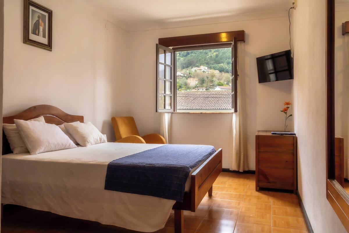 Un dormitorio con cama, una silla, cómoda de madera, televisor y ventana con vista a un pueblo.