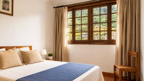 A bedroom with a large bed, a wooden chair, a window with curtains, and a small table with a lamp and plant.