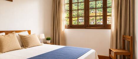 Une chambre avec un grand lit, une chaise en bois, une fenêtre avec des rideaux et une petite table sur laquelle se trouvent une lampe et une plante.