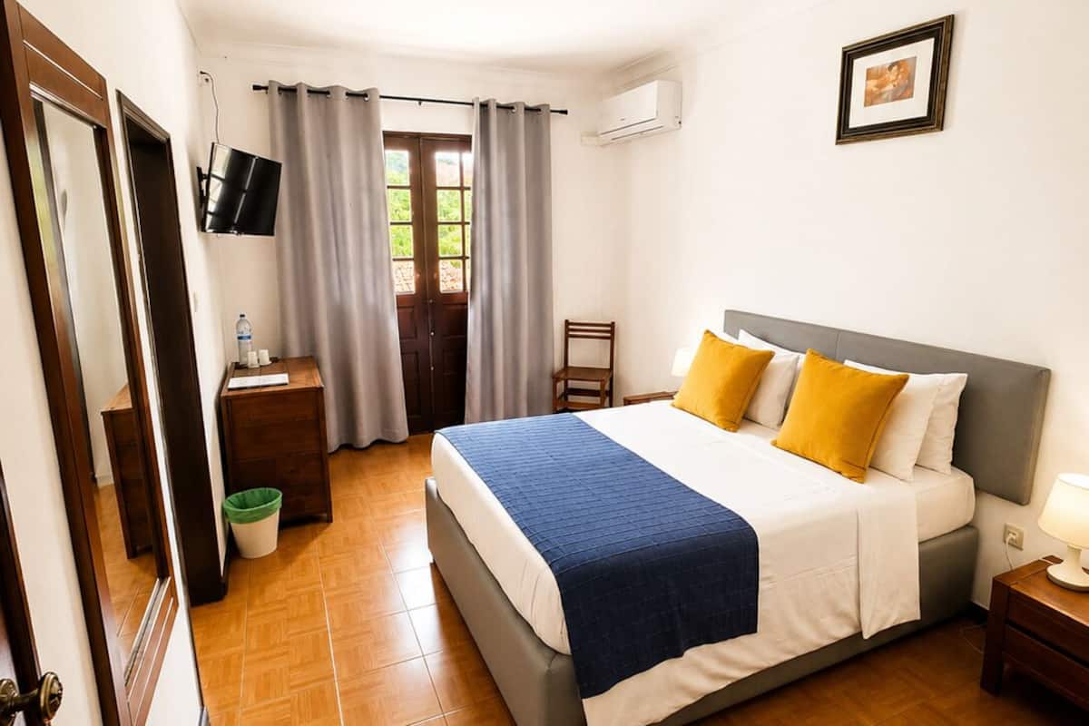 Habitación de hotel con cama, mesita de noche, una silla, televisor montado en la pared y ventana con cortinas.