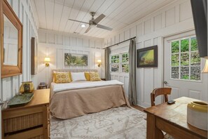 Ein Schlafzimmer mit Bett, Nachttischen, Schreibtisch, Stuhl, Deckenventilator und einem Fenster mit Vorhängen.
