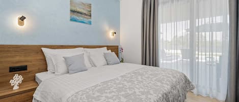 A bedroom with a wooden headboard, a bed with a patterned bedspread, two wall-mounted lights, a painting, and a window with sheer curtains.
