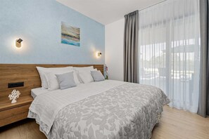 Ein Schlafzimmer mit einem hölzernen Kopfteil, einem Bett mit einem gemusterten Bettbezug, zwei Wandleuchten, einem Gemälde und einem Fenster mit transparenten Vorhängen.