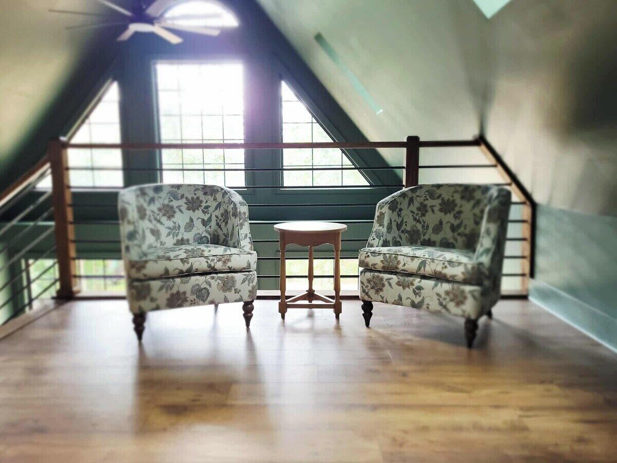 Quarto com duas poltronas estampadas, uma mesa de madeira pequena e vista para uma escada e janelas.