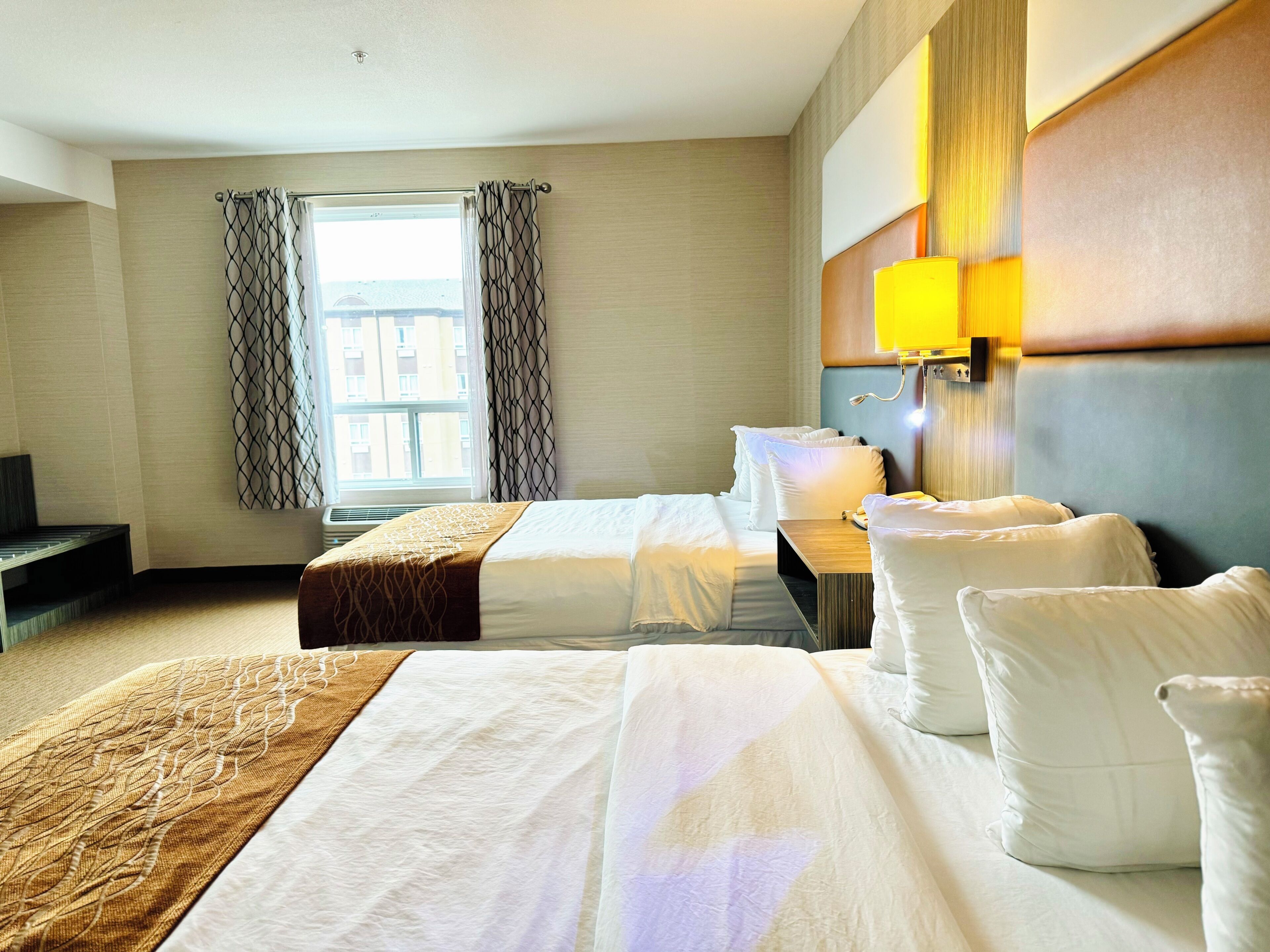 A hotel room with two beds, a bedside table, a lamp, and a window with curtains.