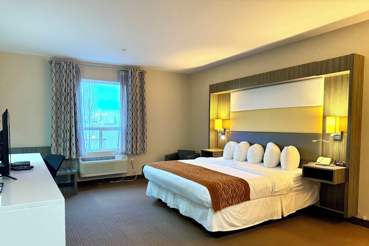 Habitación de hotel con cama, mesitas de noche, escritorio, silla, televisor, ventana con cortinas y lámpara de pared.