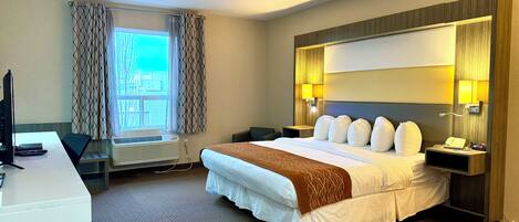 A hotel room with a bed, bedside tables, a desk, a chair, a TV, a window with curtains, and a wall-mounted lamp.