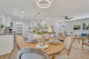Eine moderne Küche mit weißen Schränken, eine Essecke mit einem Holztisch und ein Wohnzimmer mit Sofa und Deckenventilator.
