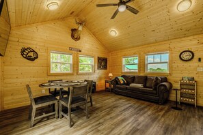 Ein Interieur einer Holzhütte mit Esstisch, Stühlen, einer Couch, einem Deckenventilator und einem aufgehängten Hirschkopf.