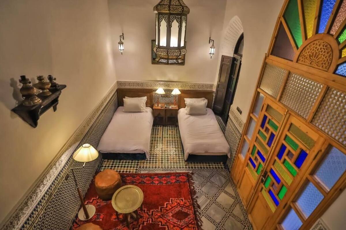 A room with two beds, a patterned floor, a decorative wall, and a small table with a lamp.