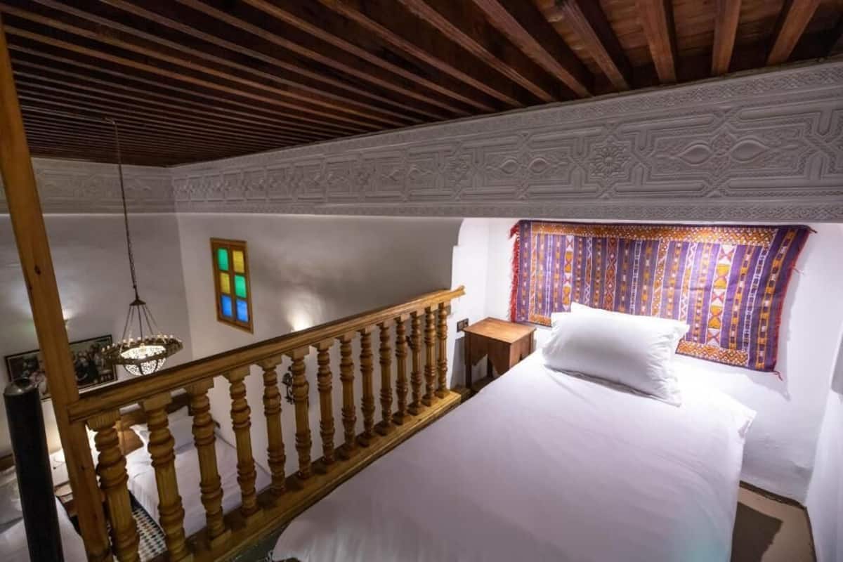 A bedroom with a wooden staircase, a bed with white bedding, a patterned curtain, and a wall-mounted light fixture.