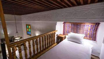 Un dormitorio con escalera de madera, cama con ropa de cama blanca, cortina estampada y lámpara fijada a la pared.