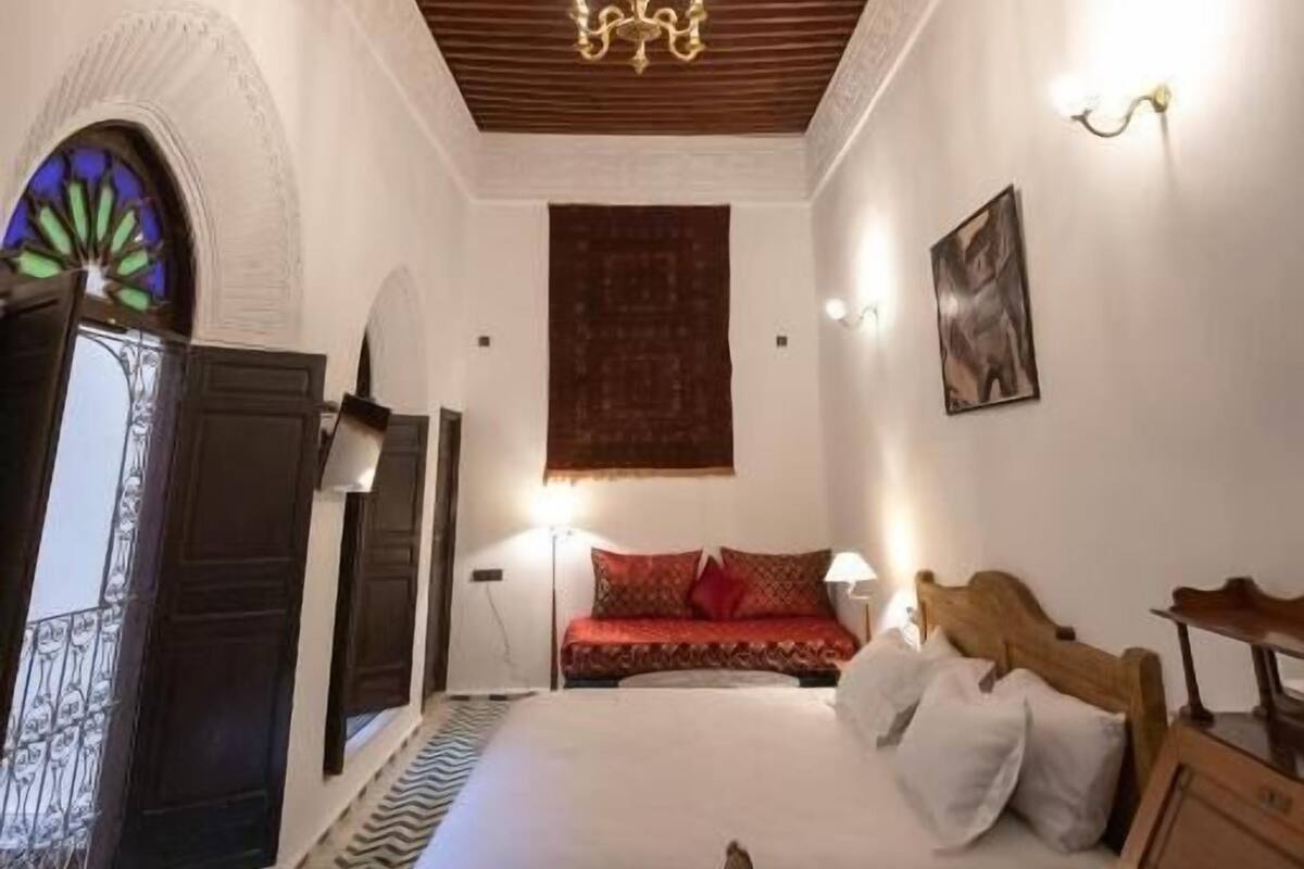 A bedroom with a wooden ceiling, a bed, a chandelier, a wall-mounted light, and a window with a view.