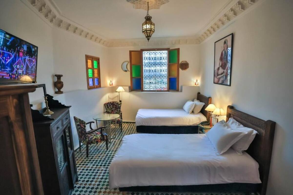 A hotel room with two beds, a television, a small table, a chair, and a decorative window.