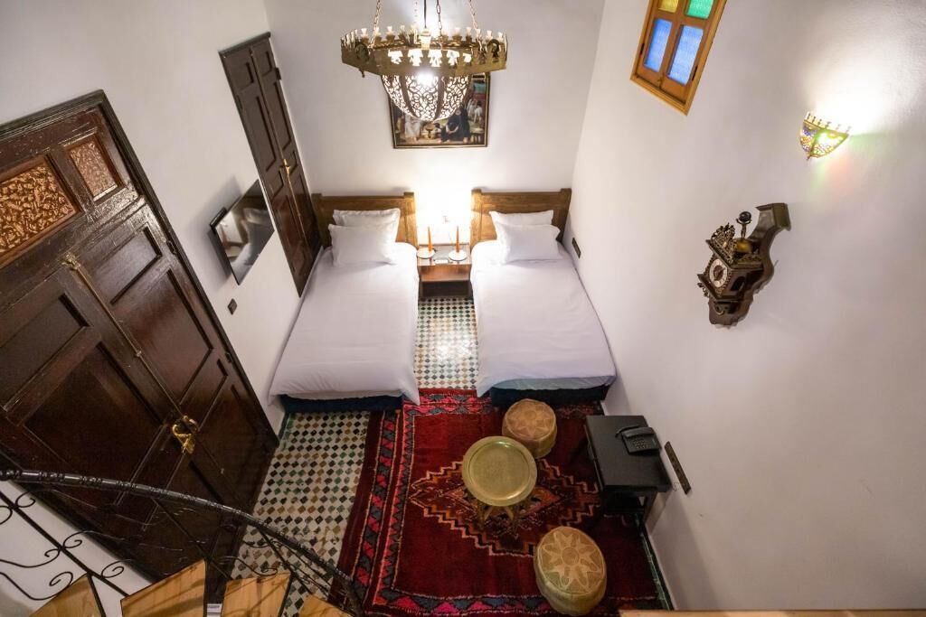 Une chambre avec deux lits, un lustre, un tapis à motifs, une petite table et une horloge murale.