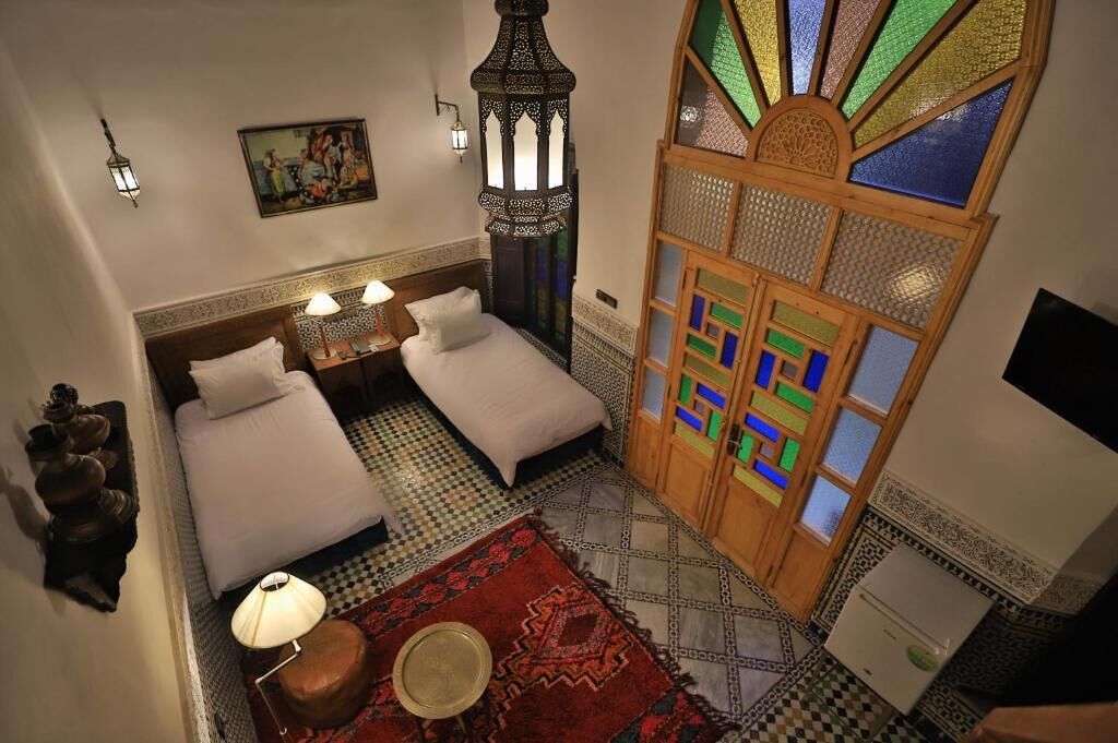 A room with two beds, a nightstand, a lamp, a decorative lantern, a wall-mounted picture, and a colorful window.