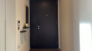 A narrow hallway with a dark wooden door, a small shelf with a mirror and a potted plant, and a light switch on the wall. - The Brompton Residence (London)