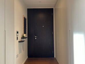 A narrow hallway with a dark wooden door, a small shelf with a mirror and a potted plant, and a light switch on the wall. - The Brompton Residence (London)