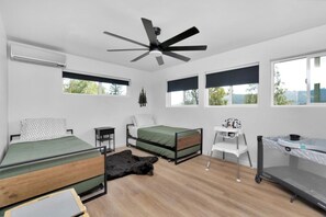A modern bedroom with two beds, a desk, a chair, a ceiling fan, and large windows.