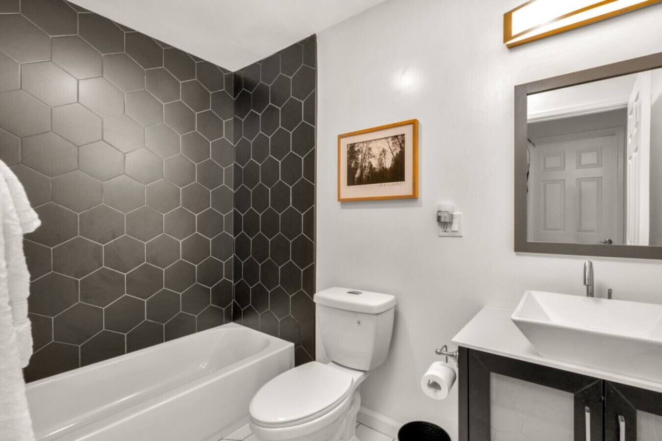 Uma casa de banho com parede de azulejos hexagonais, uma sanita, uma banheira, uma pia e um quadro emoldurado na parede.