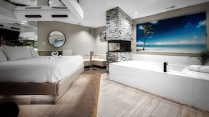 A modern bedroom with a large bed, a round mirror, a stone fireplace, and a view of the beach. - Sybaris Pool Suites Frankfort (Frankfort)