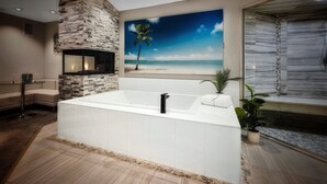 A modern bathroom with a large bathtub, a stone accent wall, a painting of a beach, and a potted plant. - Sybaris Pool Suites Frankfort (Frankfort)
