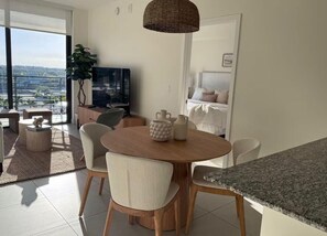 Ein modernes Wohnzimmer mit einem runden Holztisch, zwei Stühlen, einem Fernseher und einem großen Fenster mit Blick auf eine Stadtlandschaft.
