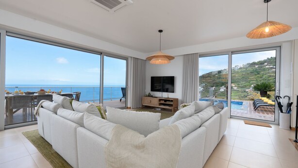 Een ruime woonkamer met een grote bank, een televisie en een eethoek met uitzicht op zee.