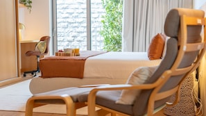 Ein modernes Hotelzimmer mit einem großen Bett, einem Sessel, einem kleinen Schreibtisch mit Lampe und einem Fenster mit Blick auf Bäume.