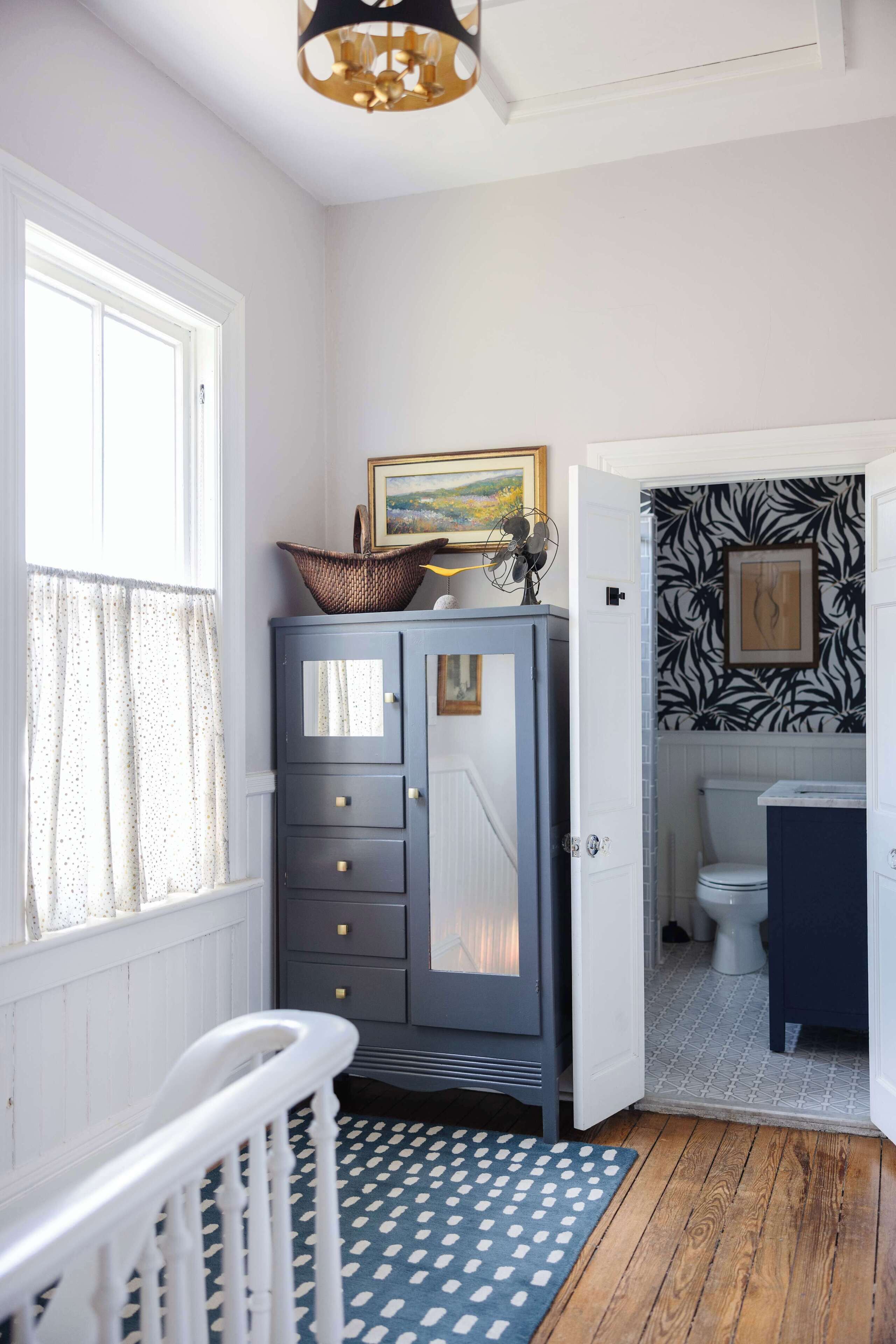 A room with a crib, a dresser with a basket and picture, a patterned wall, a toilet, and a patterned rug.