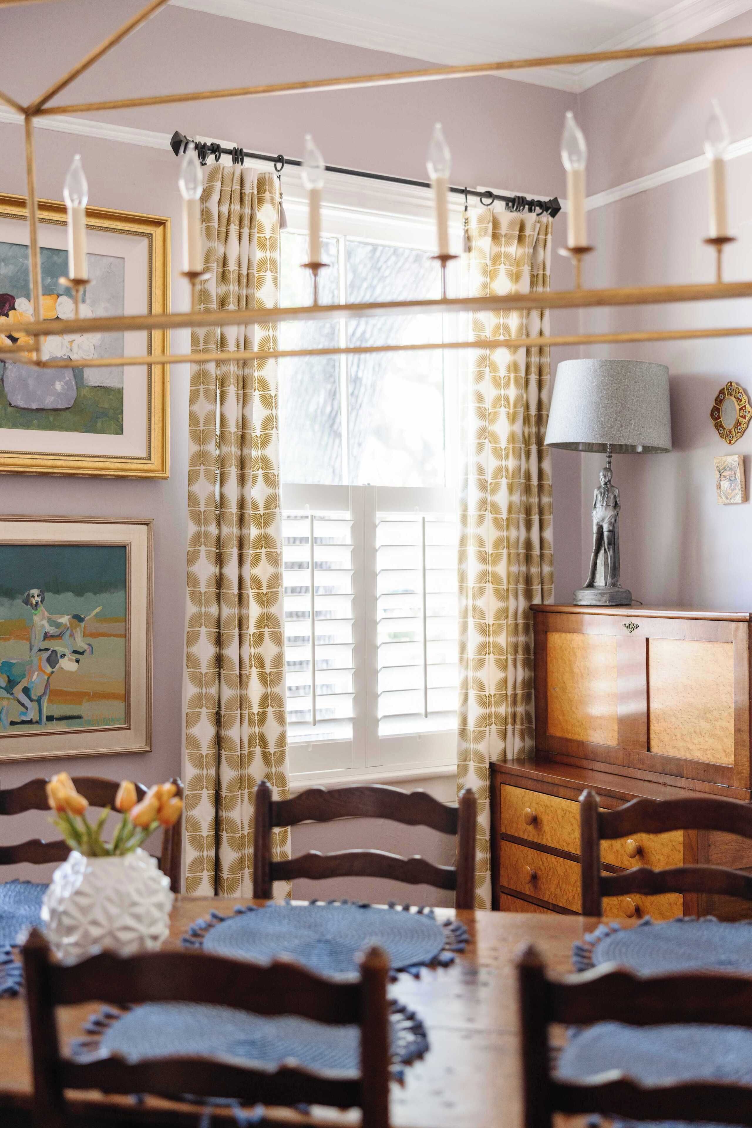 A dining area with a wooden table, chairs, a vase with flowers, a lamp, and a window with curtains.