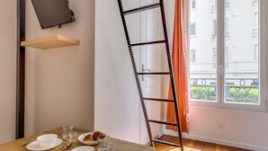 A dining area with a wooden table, white plates, and cutlery, a TV mounted on the wall, and a window with a view of buildings and greenery. - Studio 2P - Roland Garros Stadium and Bois de Boulogne (Paris)