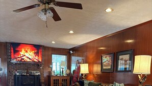 A living room with a fireplace, a large sofa, a ceiling fan, and a television displaying a fire.